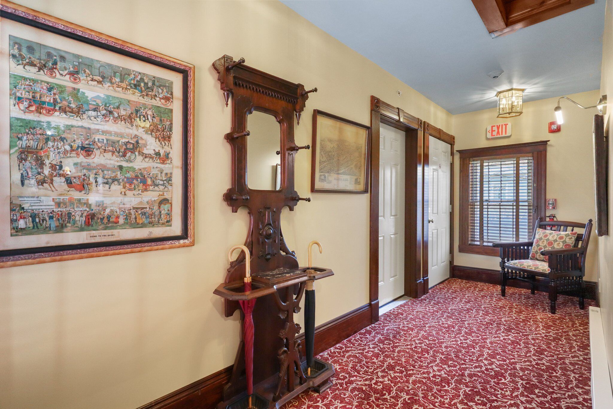 A hallway with a red patterned carpet, a large framed historical painting, a wooden mirror, and a chair with a cushion.