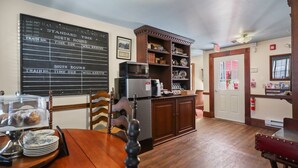 A dining area with a wooden table, chairs, a refrigerator, and a microwave. There is a blackboard with train schedules on the wall.
