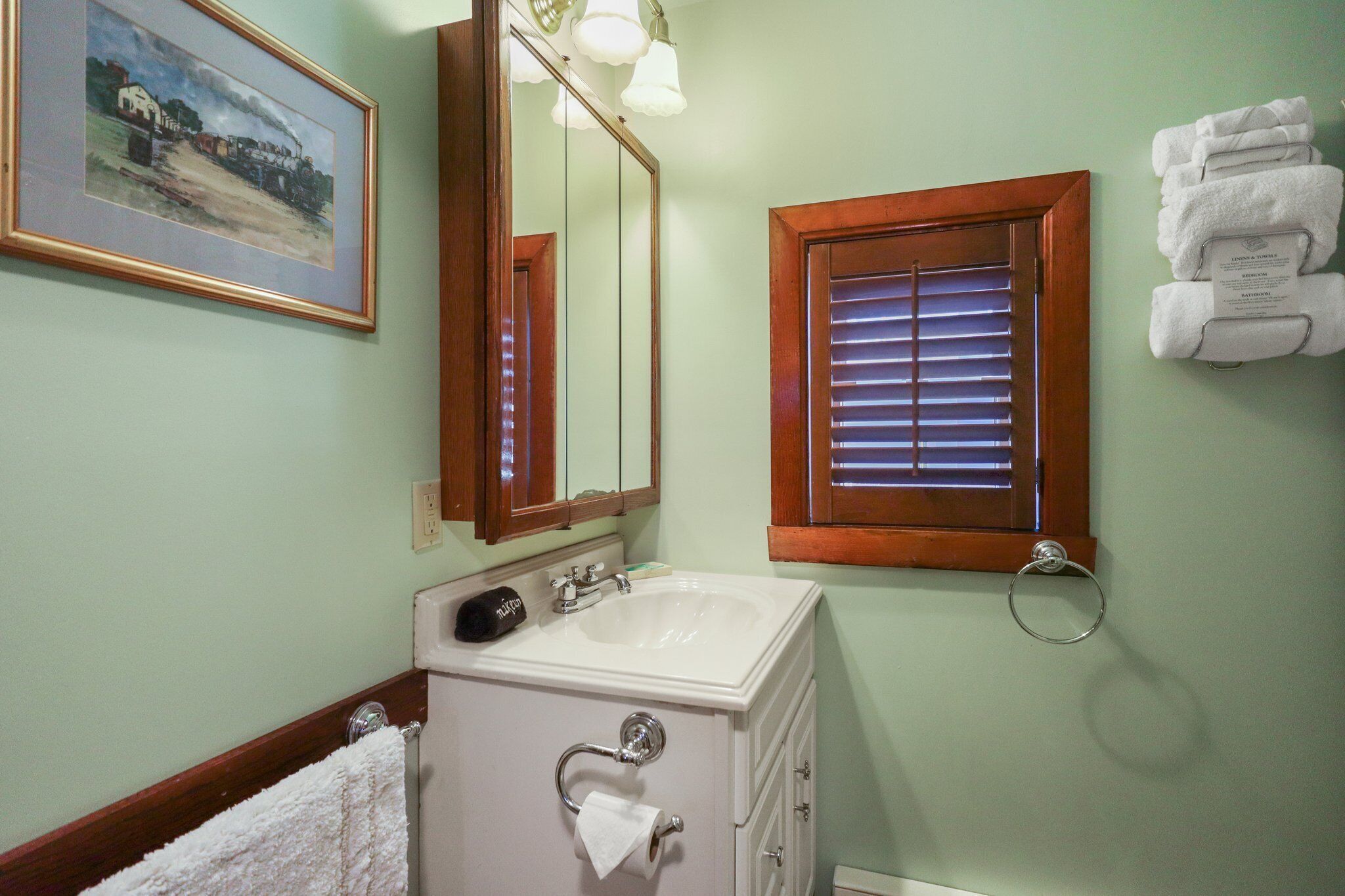 A bathroom with a white sink, a mirror, a framed painting, and a window with blinds.
