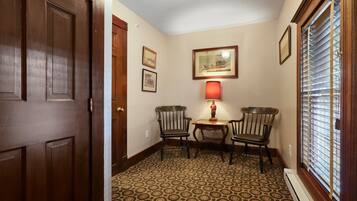A hallway with patterned carpet, wooden doors, two chairs, a small table with a lamp, and a window with blinds.