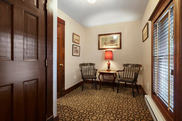 A hallway with patterned carpet, wooden doors, two chairs, a small table with a lamp, and a window with blinds.