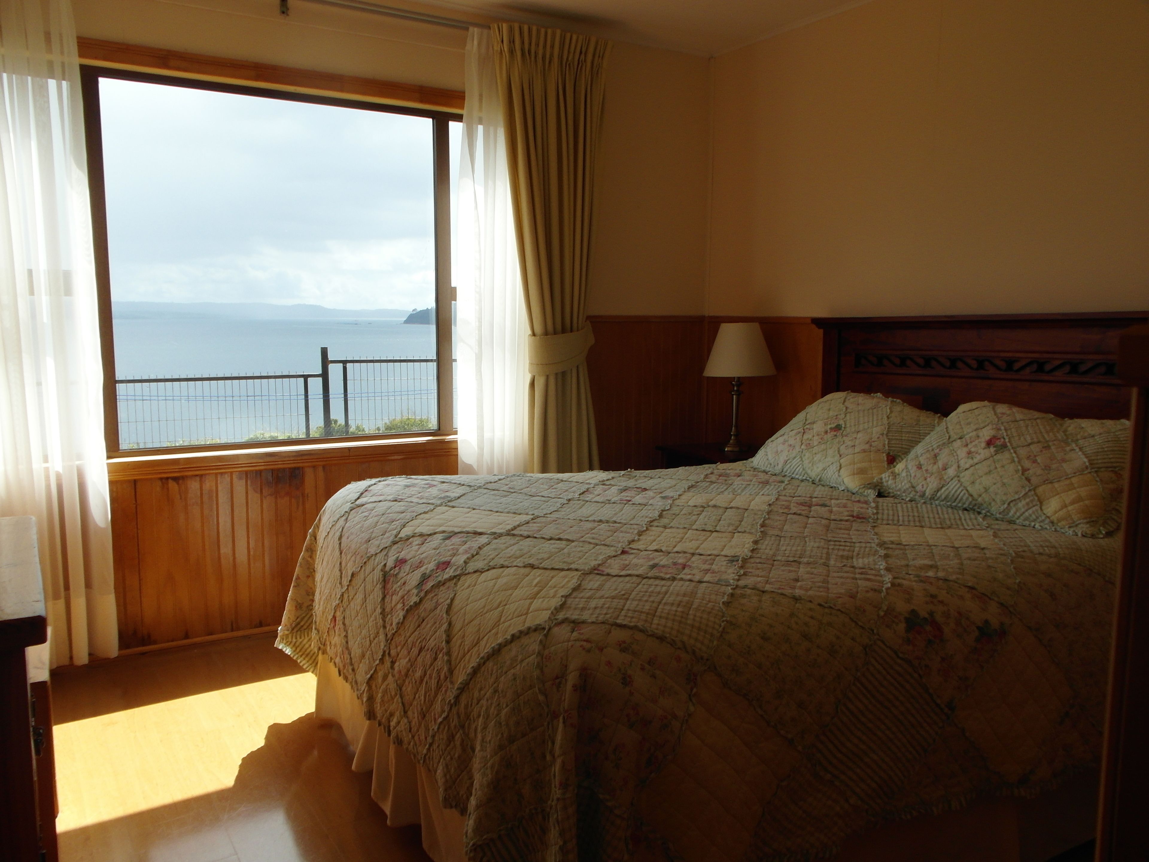 A bedroom with a large window offering a view of the sea, a bed with a floral quilt, and a wooden headboard.