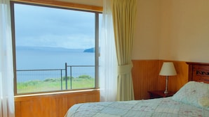 Une chambre avec une grande fenêtre, un lit recouvert d’une courtepointe à motifs floraux, une table de chevet avec une lampe, et une vue sur la mer.