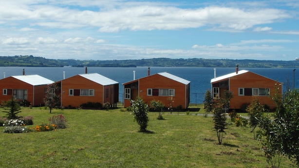 Une rangée de trois cabanes en bois aux toits blancs, se dressant sur un fond composé d’une vaste étendue d’eau et de collines lointaines.