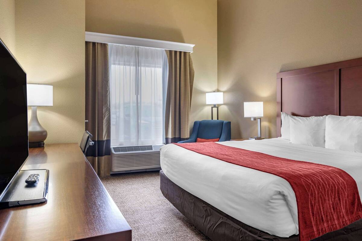 Quarto de hotel com uma cama grande, uma escrivaninha com controle remoto, duas luminárias de cabeceira, uma cadeira e uma janela com cortinas.