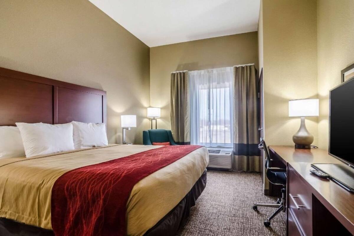 Quarto de hotel com uma cama grande, uma escrivaninha com televisão, uma cadeira e uma janela com cortinas.