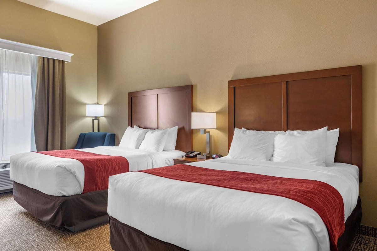 Quarto de hotel com duas camas, cada uma com roupa de cama branca e mantas vermelhas. Há duas mesas de cabeceira com luminárias, uma cadeira e uma janela com cortinas.