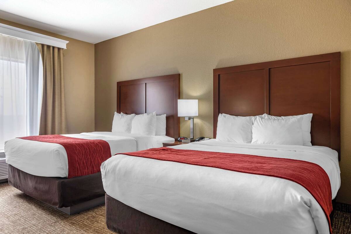 Quarto de hotel com duas camas, uma mesa de cabeceira, uma luminária e uma janela com cortinas.