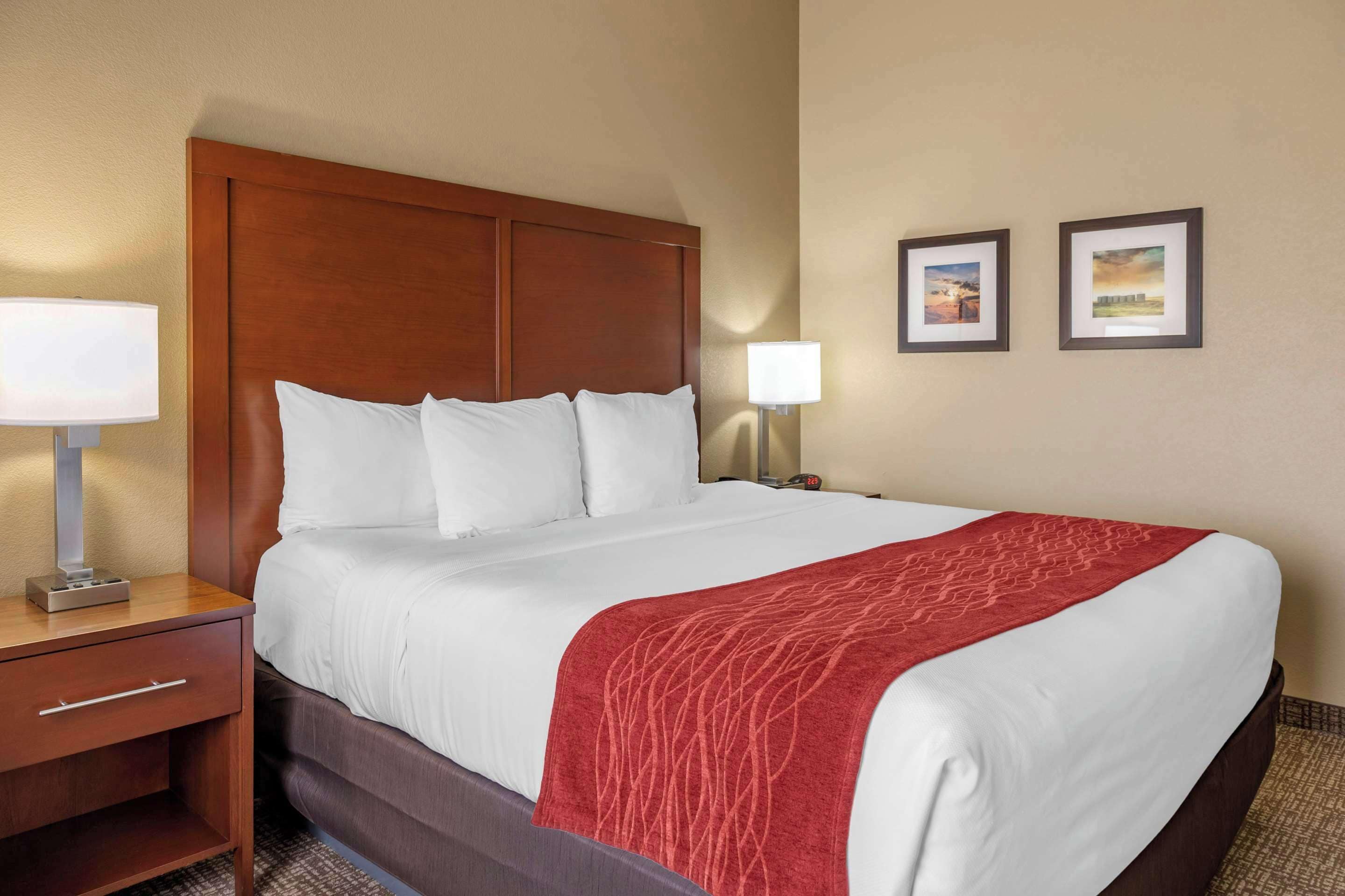 A hotel room with a large bed, a wooden headboard, a nightstand with a lamp, and two framed pictures on the wall.