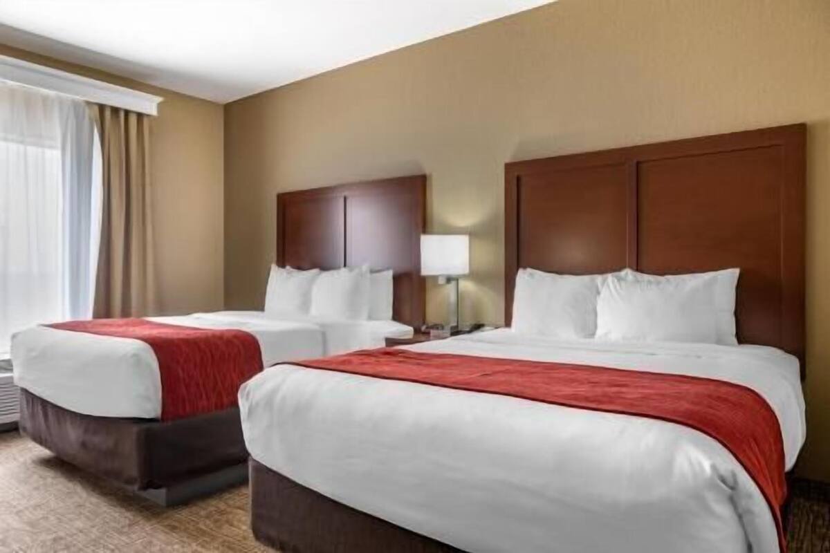 Quarto de hotel com duas camas, uma janela grande com cortinas e cabeceira de madeira.