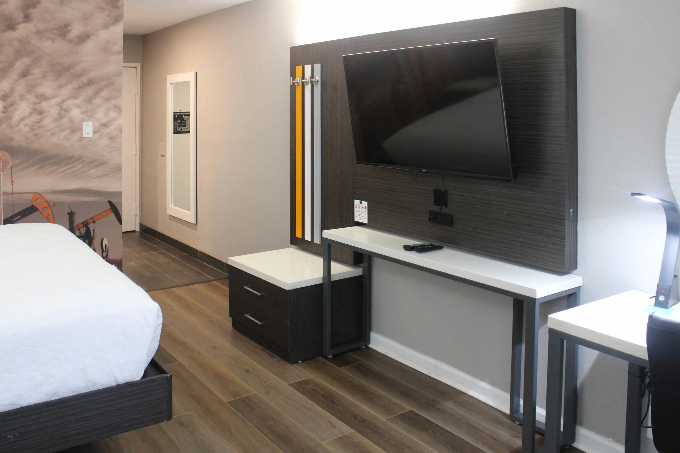 Una habitación de hotel moderna con un televisor de pantalla plana grande montado en una pared de paneles de madera, una mesita de noche blanca, un aparador de madera oscura y una cama grande con un edredón blanco.