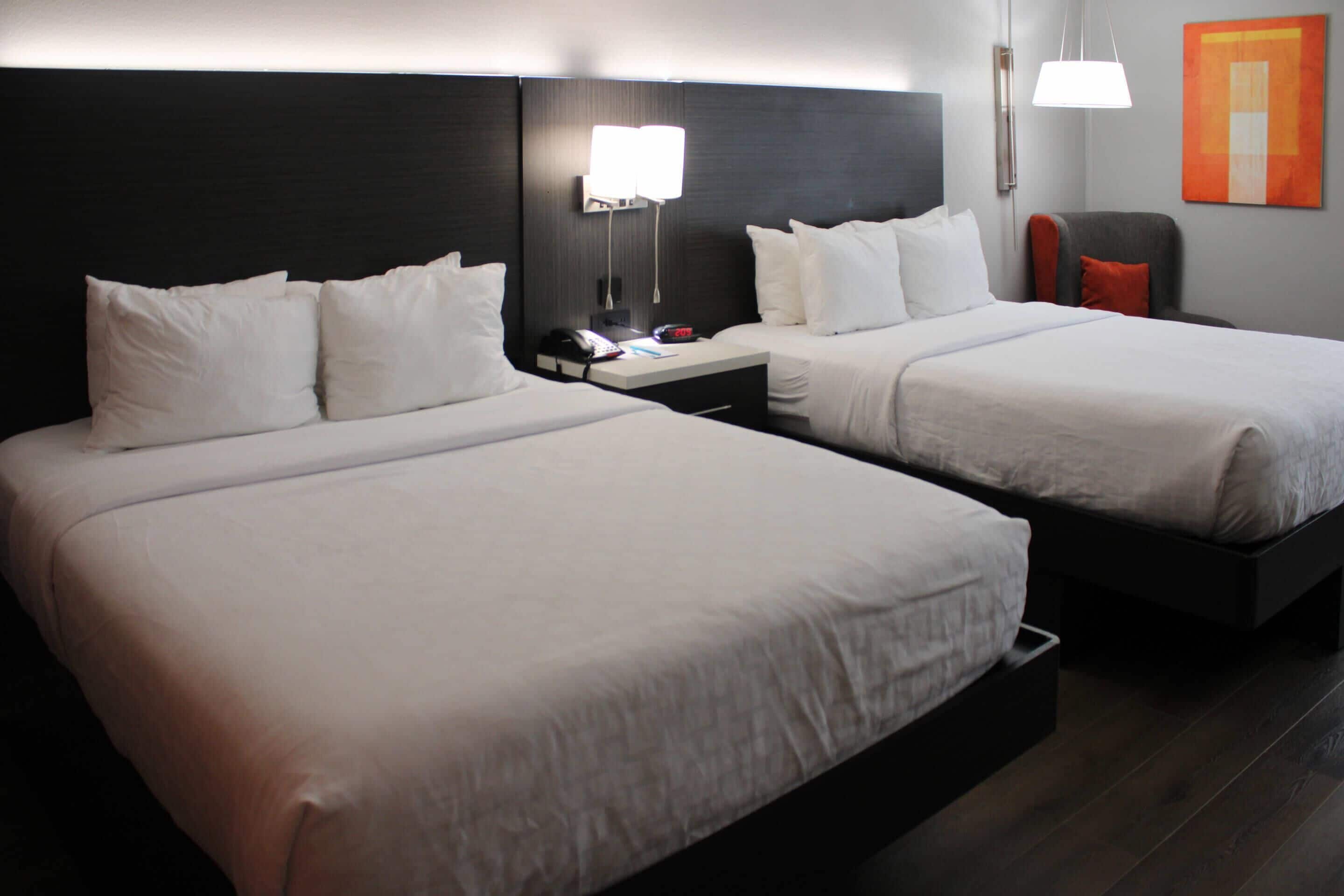 A hotel room with two beds, a nightstand with a phone and remote, a chair, and a wall-mounted lamp.