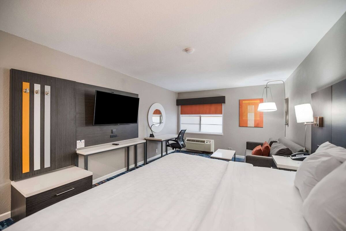 A hotel room with a large bed, a flat-screen TV, a desk with a chair, a round mirror, and a window with blinds.