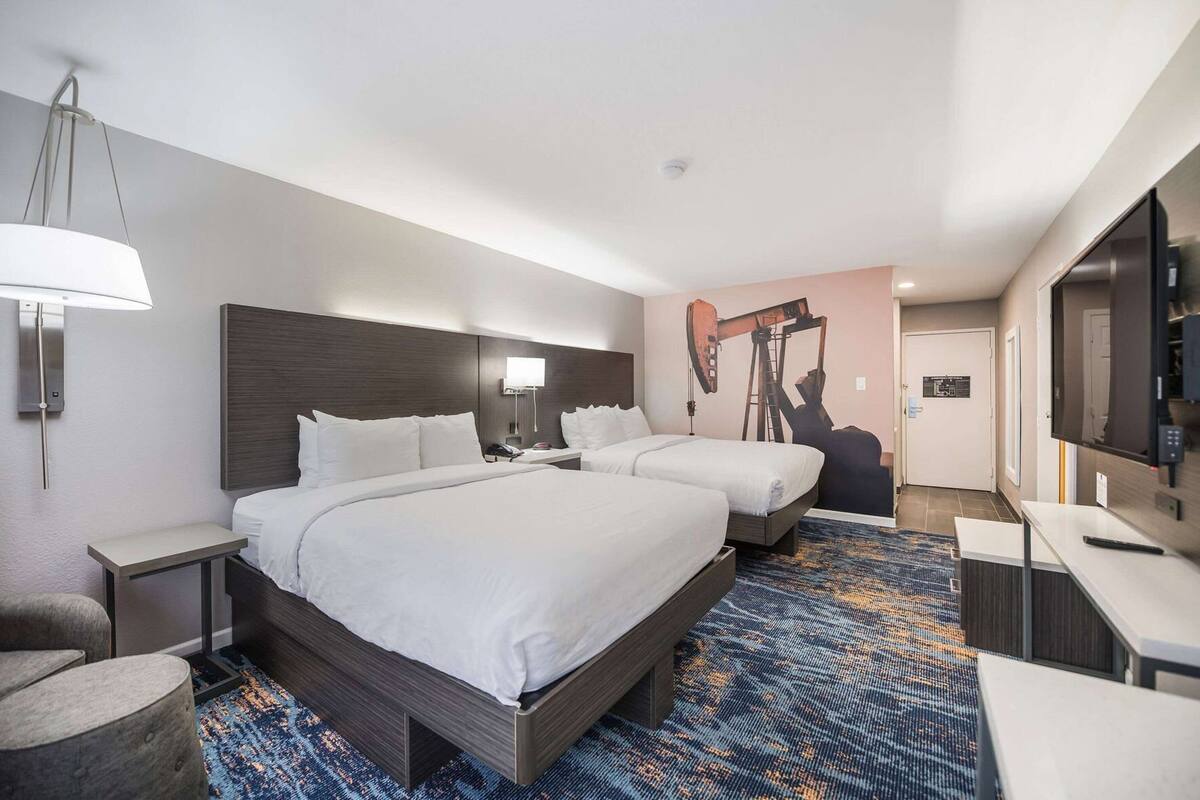 A hotel room with two beds, a desk, a chair, a lamp, and a wall-mounted TV.