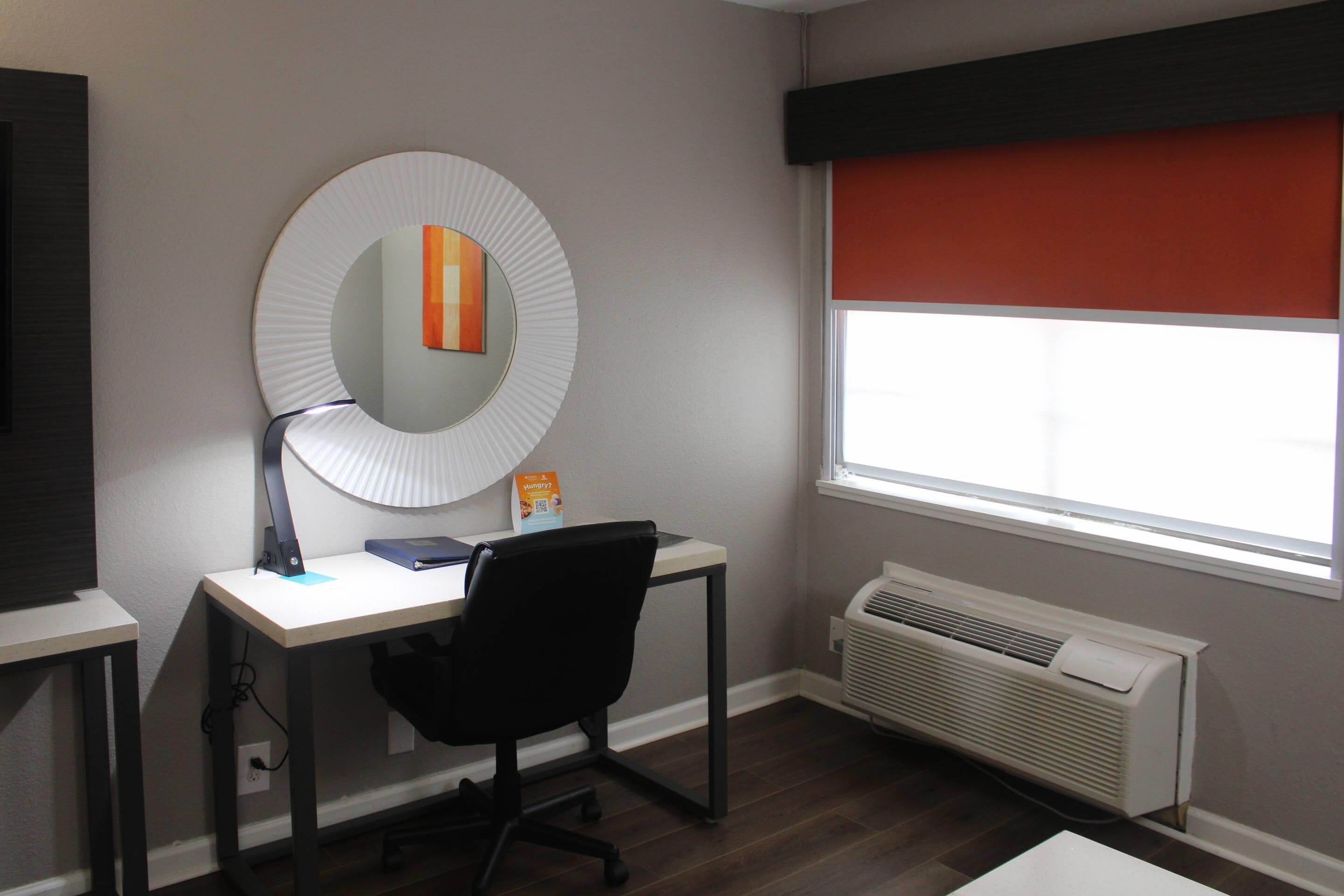 Una habitación con escritorio, silla y un espejo grande. Hay una ventana con persiana roja y un aire acondicionado de pared.
