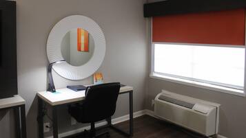 Una habitación con escritorio, silla y un espejo grande. Hay una ventana con persiana roja y un aire acondicionado de pared.