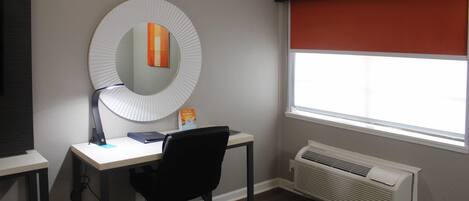 Una habitación con escritorio, silla y un espejo grande. Hay una ventana con persiana roja y un aire acondicionado de pared.