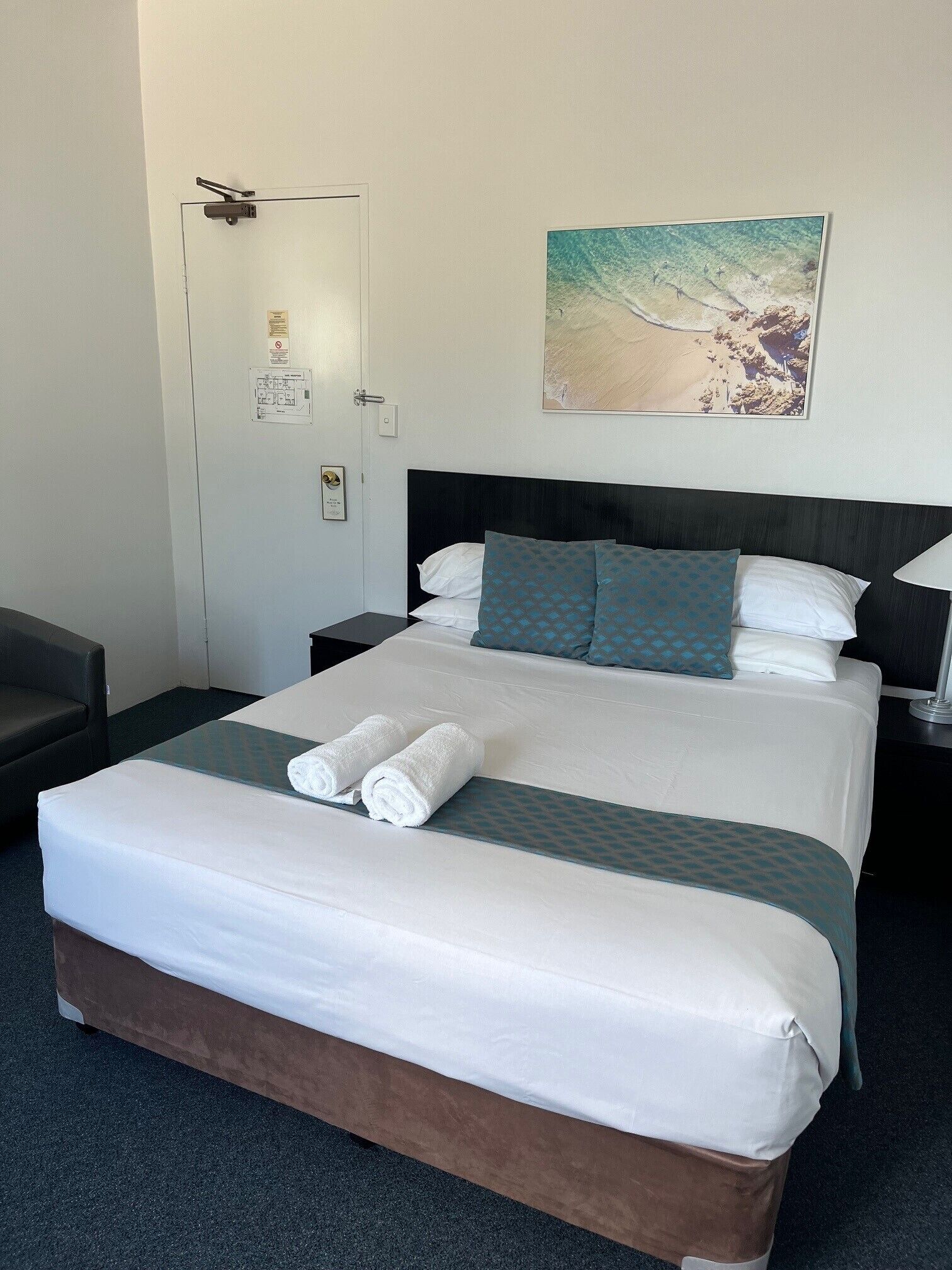 Un letto rifatto con lenzuola bianche e due asciugamani arrotolati, una testiera scura e un quadro raffigurante una spiaggia sulla parete.
