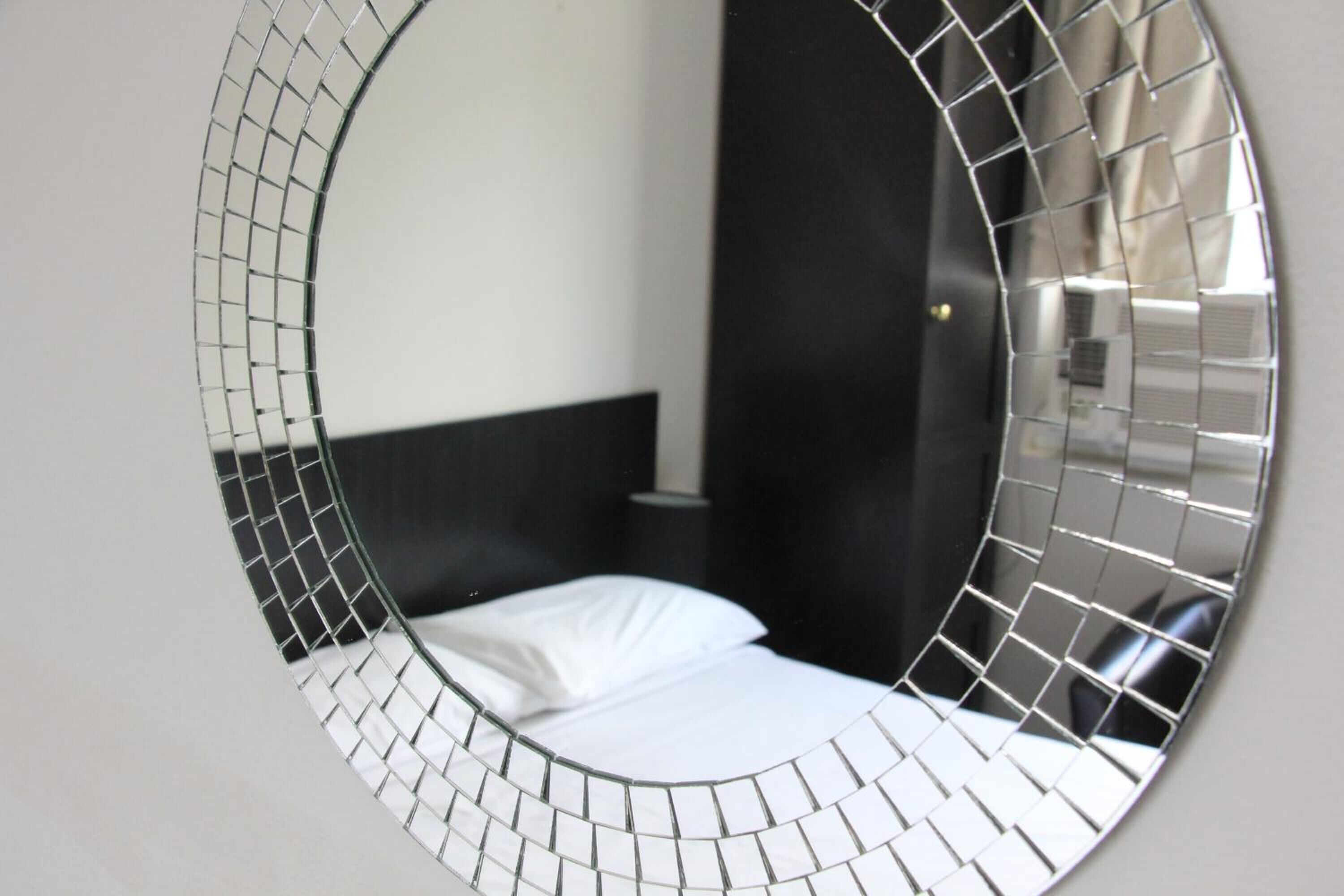 Um espelho circular com padrão em mosaico, refletindo uma cama com roupa de cama branca e cabeceira preta.