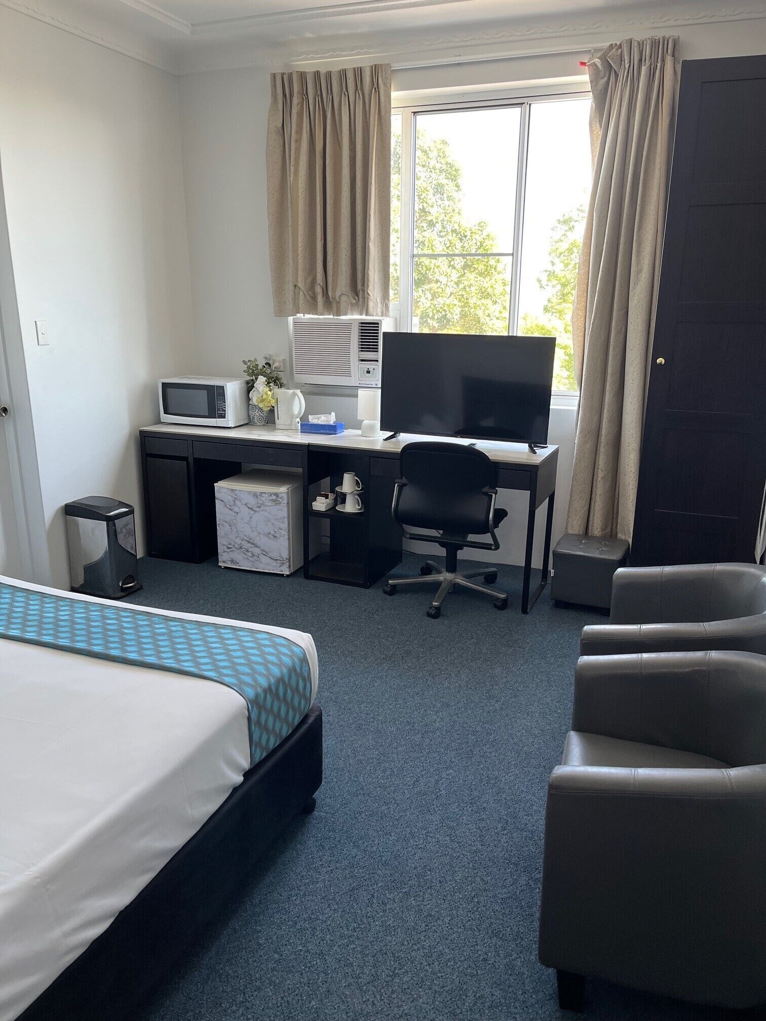 Et hotelværelse med seng, skrivebord med computer, mikroovn og et vindue med gardiner.