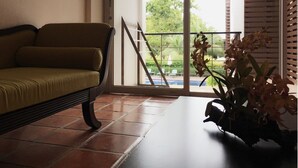 Une pièce avec un canapé, une plante en pot et une vue sur une piscine et de la verdure, visible à travers une fenêtre munie de stores.
