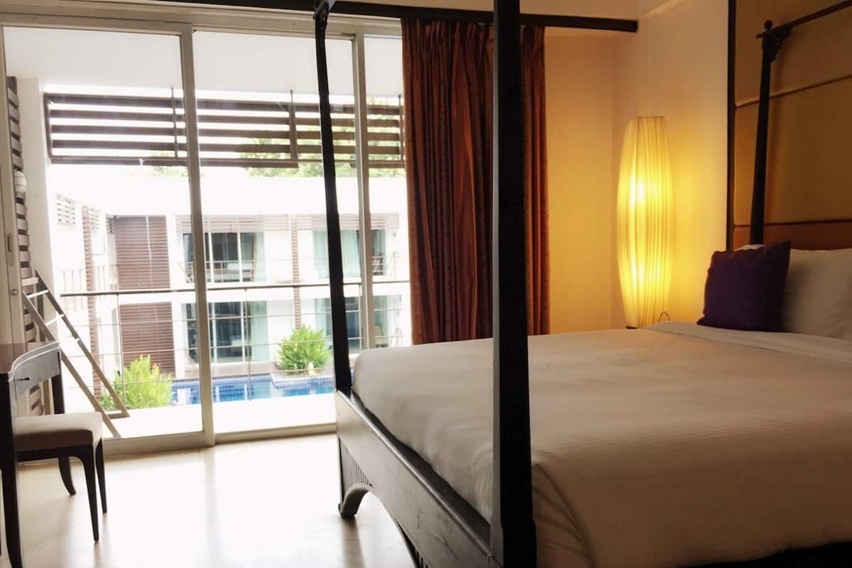 Een hotelkamer met een bed, een stoel, een raam met zonwering en uitzicht op een gebouw en een zwembad.
