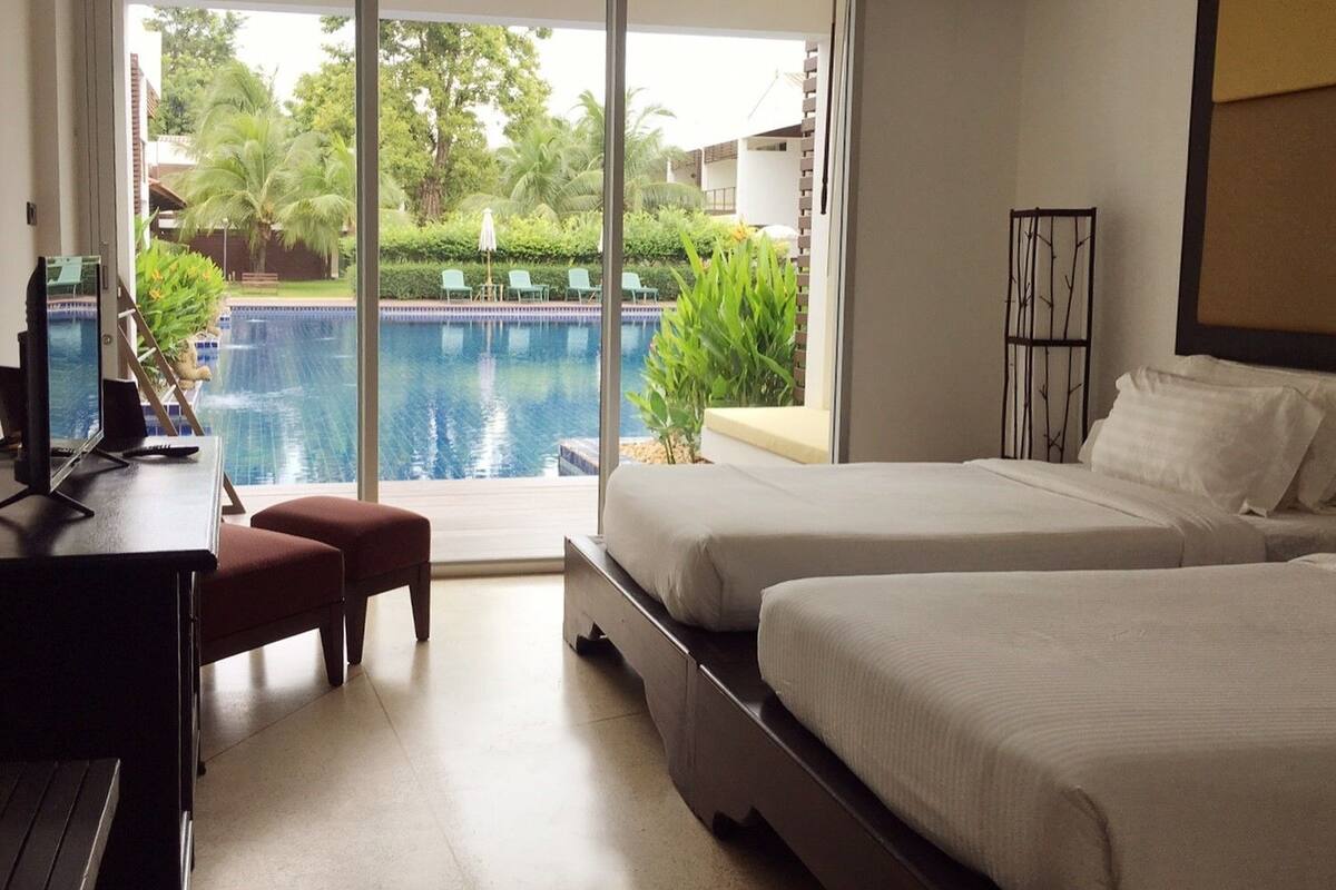 Een hotelkamer met twee bedden, een bureau en uitzicht op een zwembad en groen buiten.