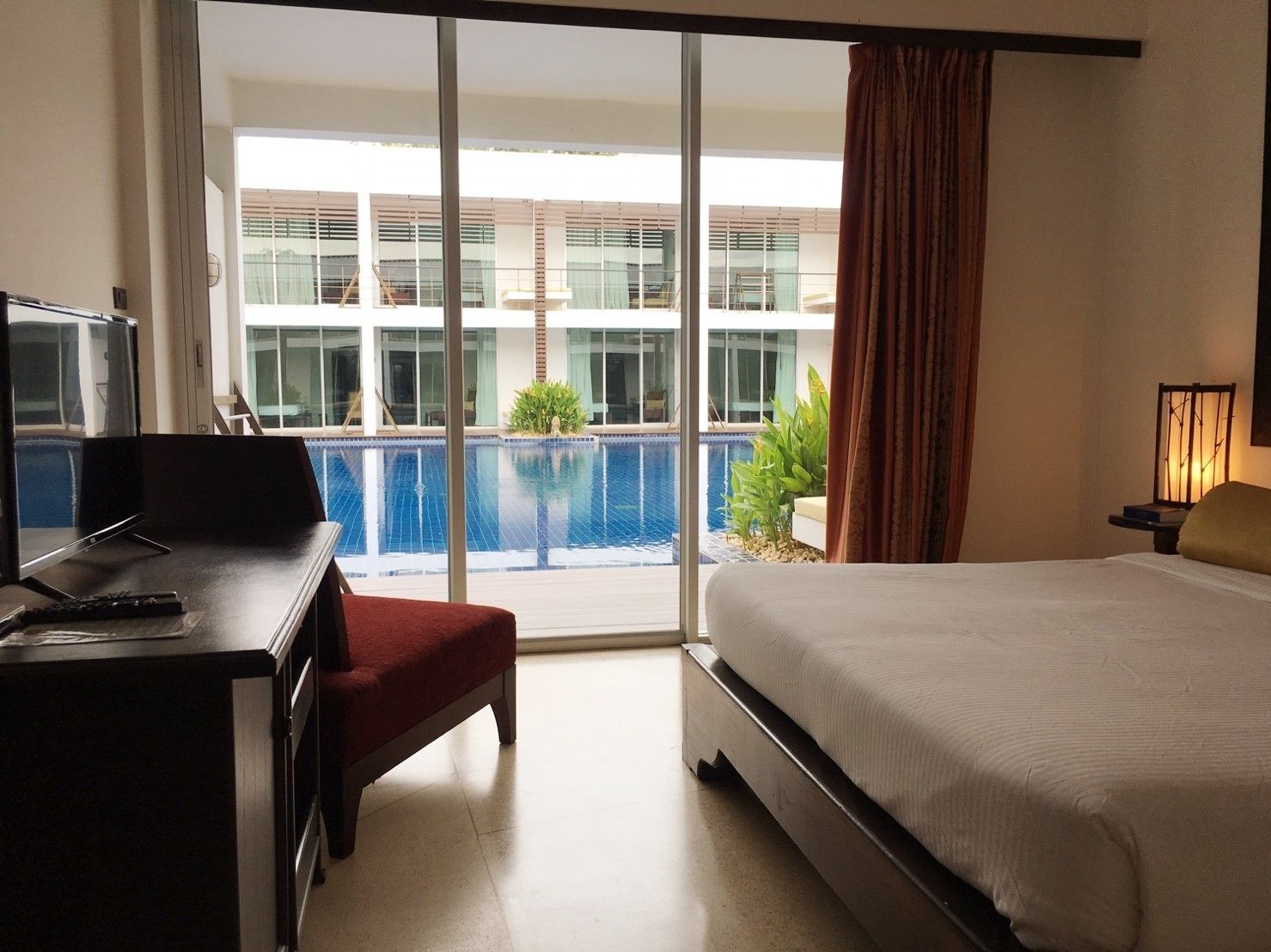 A hotel room with a bed, a desk with a TV, a red chair, a view of a swimming pool, and a balcony with curtains.