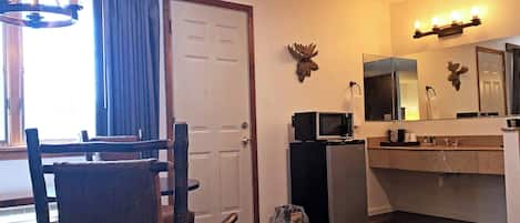 A room with a wooden chair, a door, a microwave, a refrigerator, a sink, and a mirror.