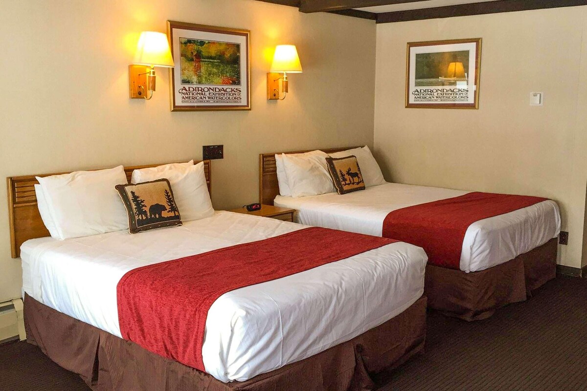 A hotel room with two beds, each with a red blanket, white linens, and a pillow with a nature design. There are wall-mounted lamps and two framed pictures of American watercolors.