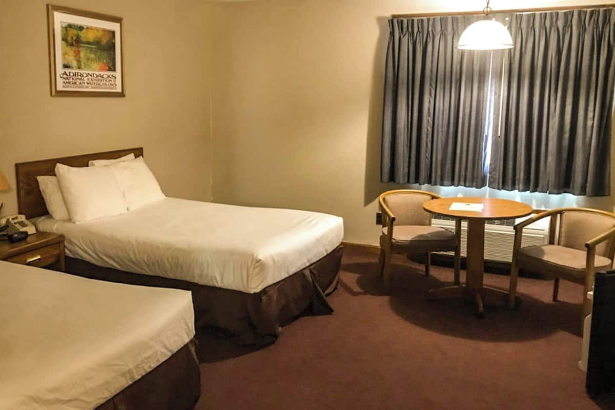 A hotel room with two beds, a desk with a lamp, a chair, a table, and a window with curtains.