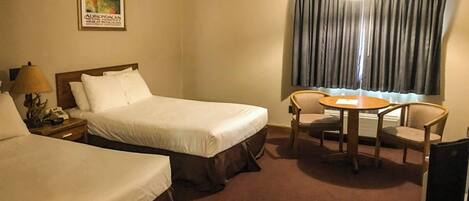 Habitación de hotel con dos camas, un escritorio con lámpara, una silla, una mesa y un ventanal con cortinas.
