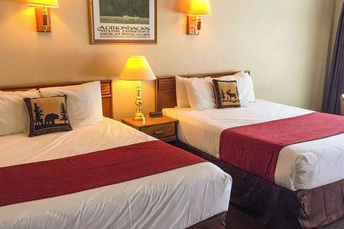 Two double beds with white linens, red throw blankets, and matching pillows. There are bedside lamps, a framed picture on the wall, and a chair in the corner.