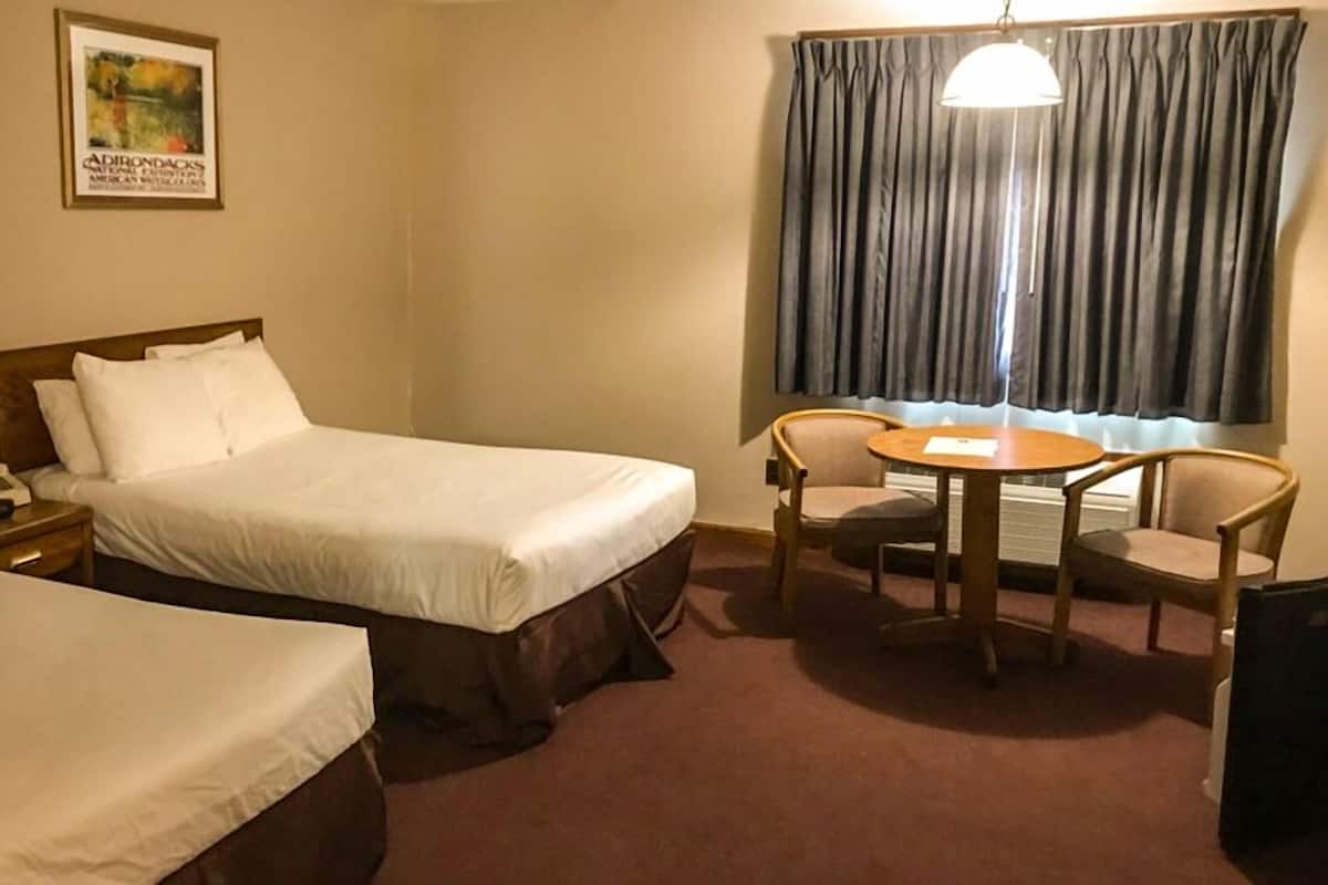 A hotel room with two beds, a table with chairs, a lamp, a telephone, and a window with curtains.