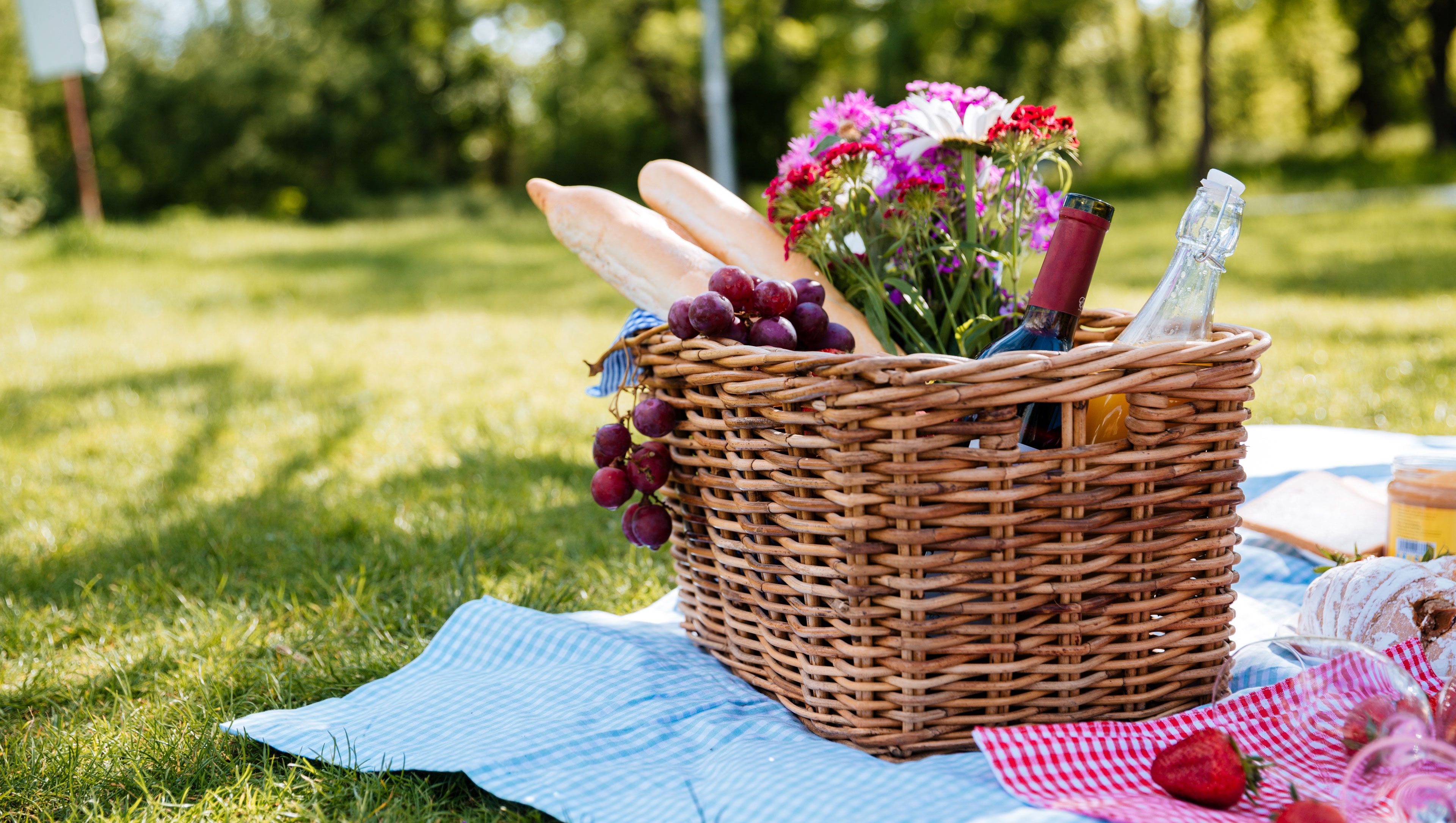 A wicker basket with a blue and white checkered cloth, containing grapes, flowers, and a bottle.