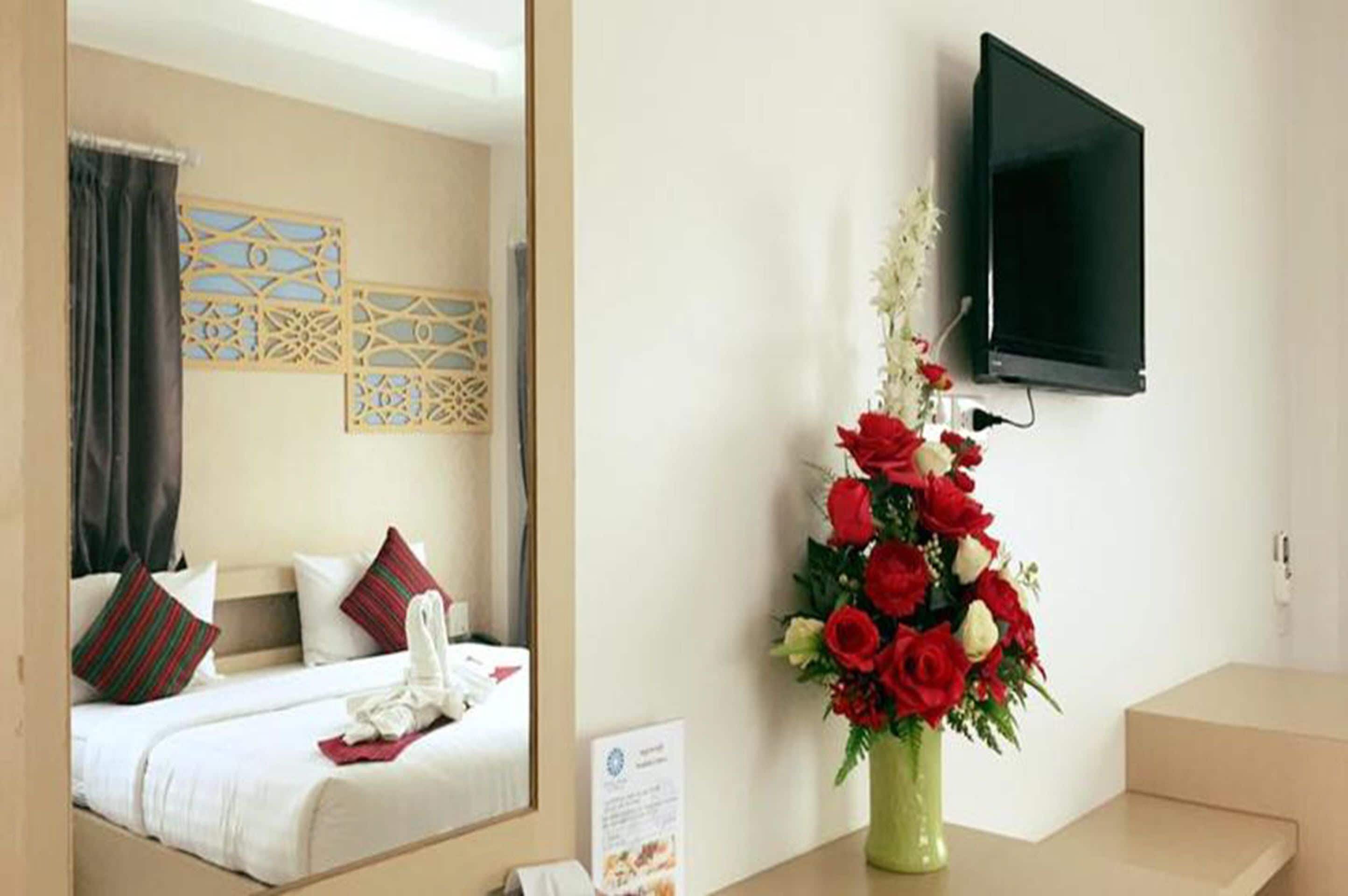 Ein Hotelzimmer mit Bett, einem an der Wand montierten Fernseher und einer Vase mit roten und weißen Blumen.