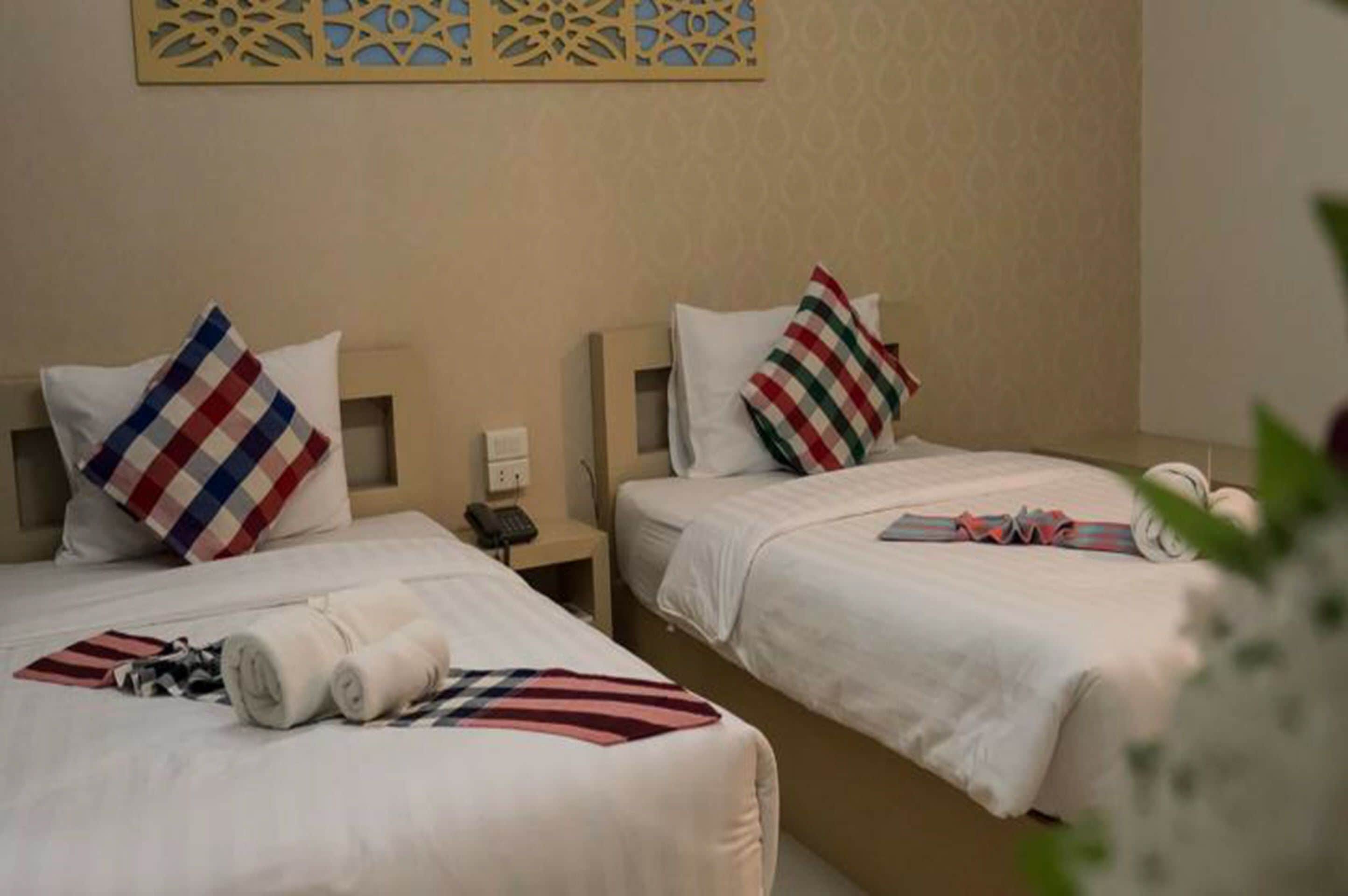 A hotel room with two beds, each with a white bedspread and a striped pillow. There is a small bedside table with a lamp and a phone.
