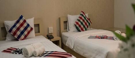 Habitación de hotel con dos camas, cada una con una colcha blanca y un cojín a rayas. Hay una mesita de noche pequeña con una lámpara y un teléfono.