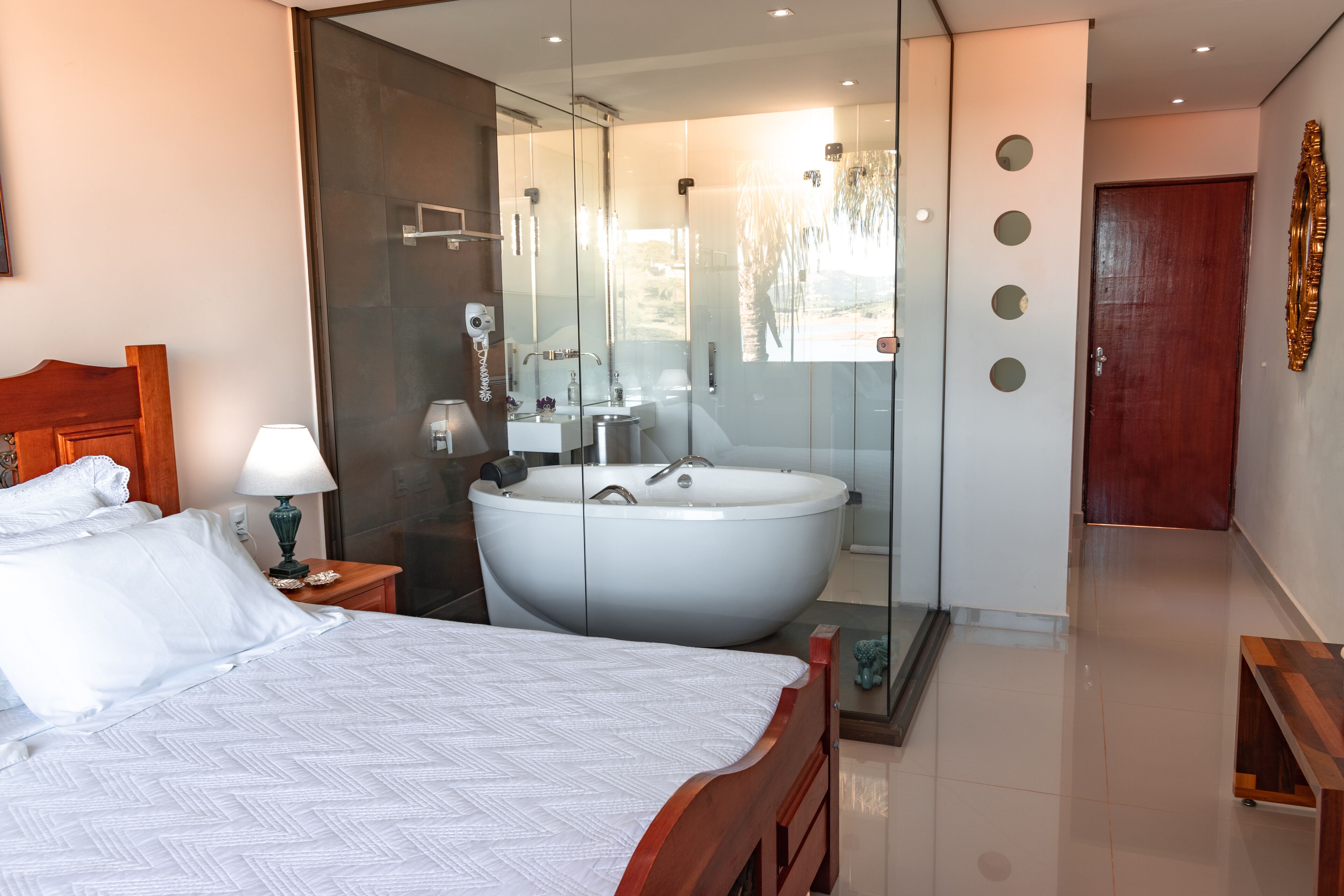 A modern hotel room with a large bed, a glass-enclosed bathroom with a freestanding bathtub, and a wooden door.