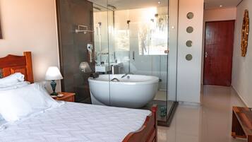 A modern hotel room with a large bed, a glass-enclosed bathroom with a freestanding bathtub, and a wooden door.