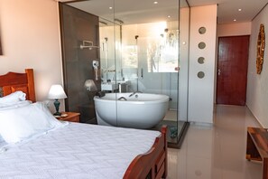 A modern hotel room with a large bed, a glass-enclosed bathroom with a freestanding bathtub, and a wooden door. - BellaMinas Pousada (Guape)