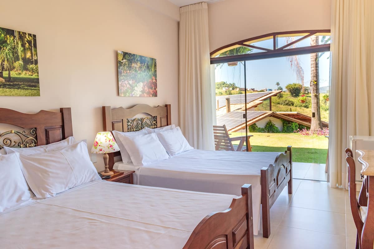 A hotel room with two beds, a nightstand with a lamp, and a large window offering a view of a garden and a pool.
