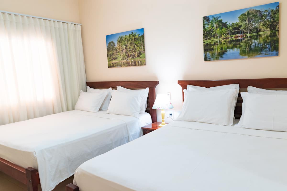 A hotel room with two single beds, a wooden headboard, a nightstand with a lamp, and two framed pictures on the wall.