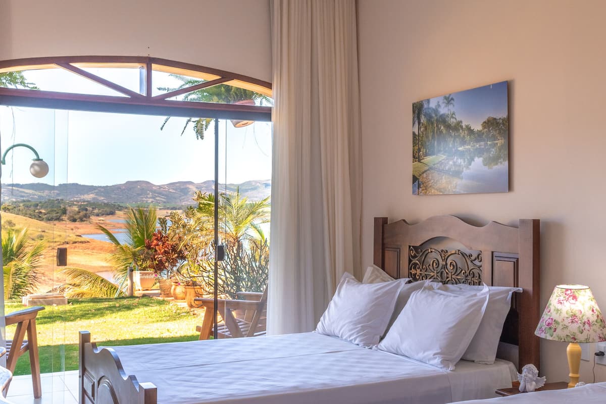 A bedroom with two beds, a large window with a view of a landscape, and a painting on the wall.