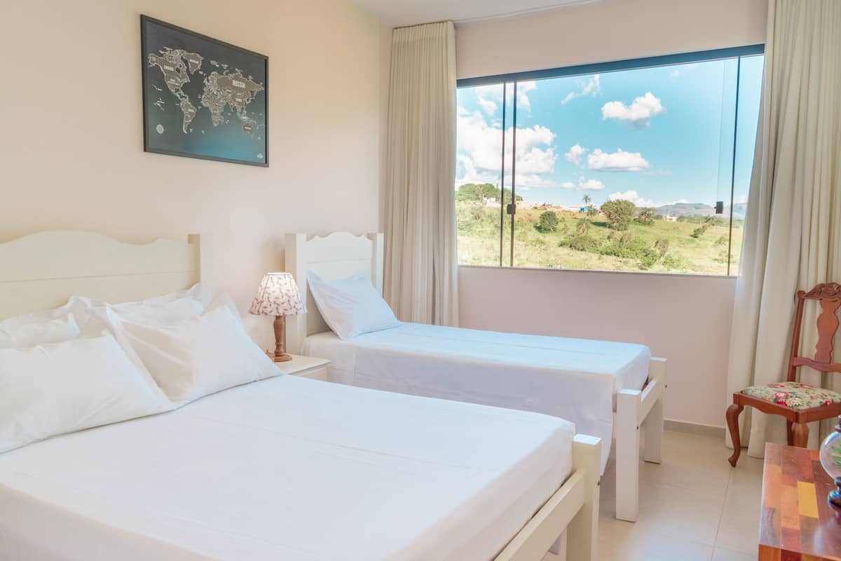 A hotel room with two beds, a world map on the wall, a large window with a view of greenery, and a wooden chair.