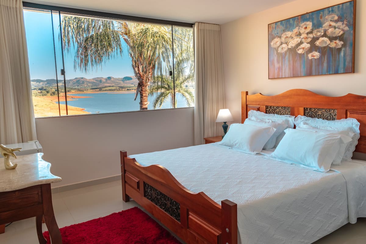 A bedroom with a large window, a bed, a nightstand, and a painting.