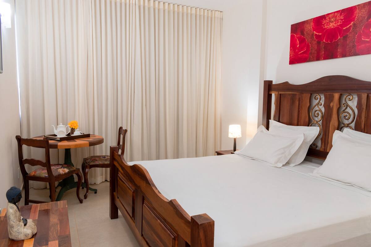 A hotel room with a wooden bed, a small table with a chair, a lamp, and a painting on the wall.