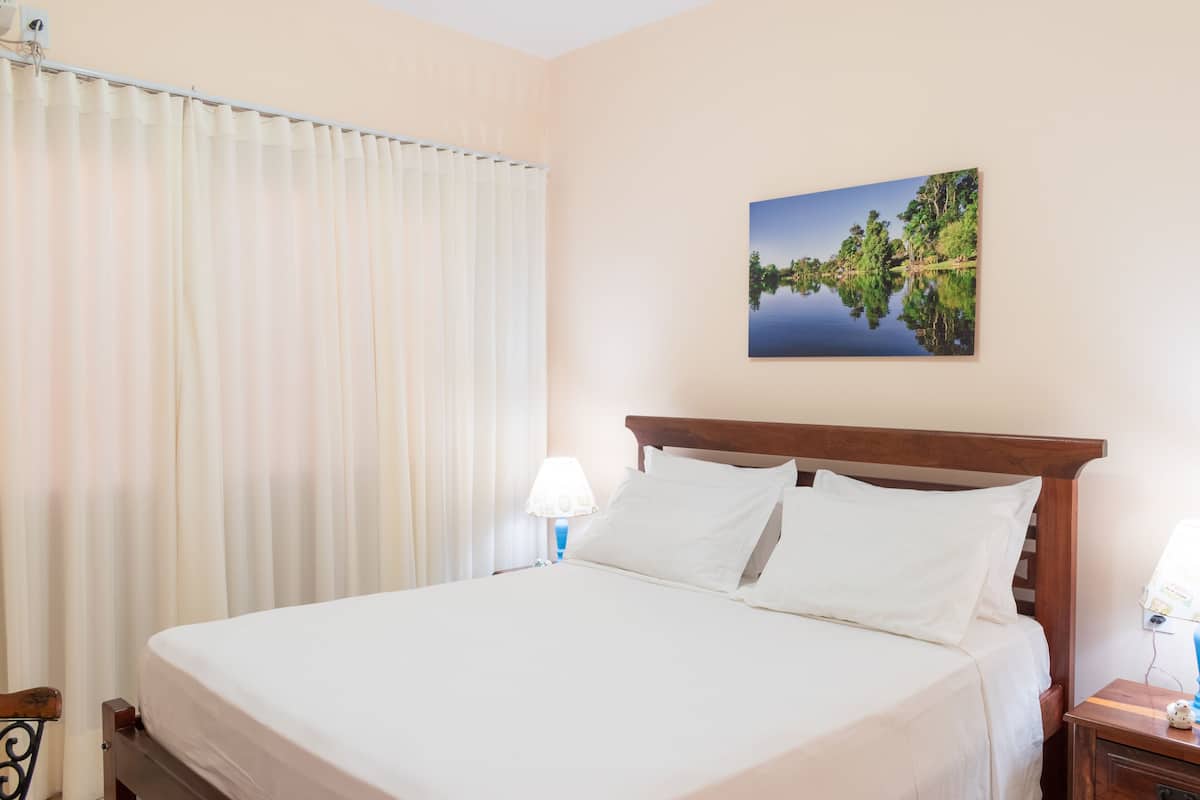 A hotel room with a large bed, white curtains, a nightstand with a lamp, and a painting of a lake on the wall.