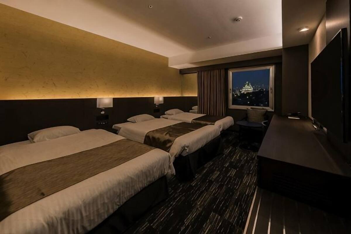 A hotel room with three beds, a large window with a city view, a desk, and a flat-screen TV.