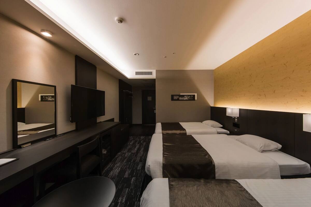 A hotel room with three beds, a desk with a chair, a TV, and a large mirror.